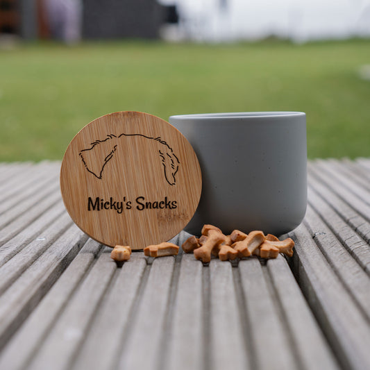 Runder Holzdeckel mit Gravur „Micky's Snacks“ und Hunde-Silhouette, daneben grauer Behälter und Hundeleckerlis auf grauer Holzterrasse. Ideal für Hundebesitzer, die Snacks für ihre Hunde aufbewahren möchten.
