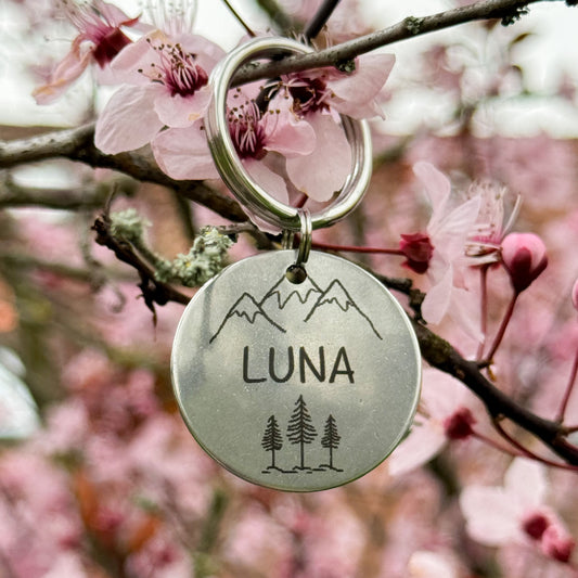 Ein runder Schlüsselanhänger mit der Gravur 'Luna' und einer Berg- und Baumillustration hängt an einem blühenden Baumzweig mit rosa Blüten.
